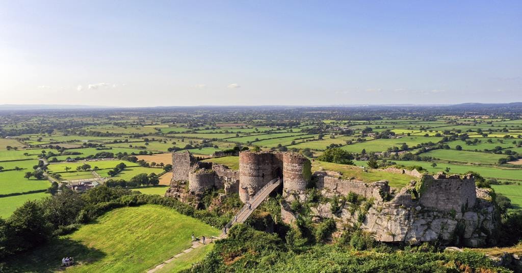 Aerial view of Beeston Castle ruins perched on a rocky hilltop, surrounded by lush green fields and patchwork countryside under a clear blue sky.